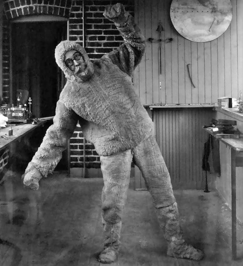 A man in glasses and a mustache strikes a dynamic pose while wearing a fluffy, full-body monster or yeti suit.