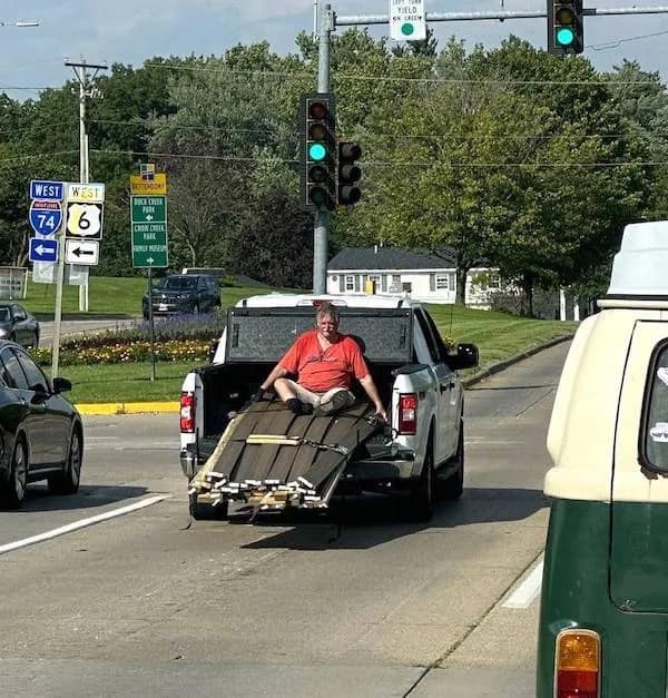 A meme dump photo of a man sitting on cargo in the back of a moving pickup truck.