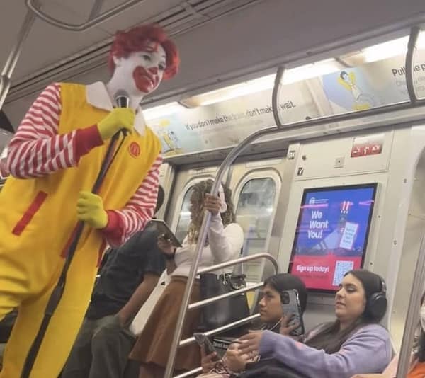 A strange and slightly threatening photo of a person who is in a full Ronald McDonald costume and is standing in a crowded subway car while holding a microphone, as if they are about to perform.