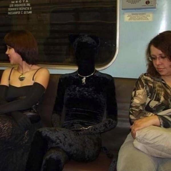 A person on a subway train who is wearing a full, head-to-toe black velvet catsuit that completely covers their face, making them look like a mysterious shadow creature with cat ears.