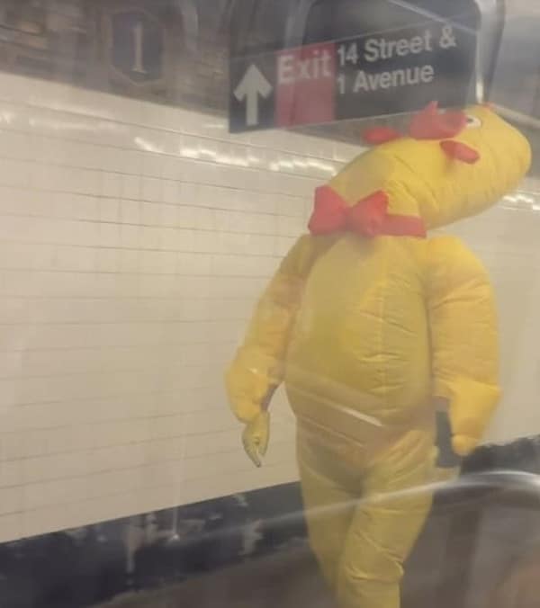 A funny photo of a person who is wearing a large, yellow, inflatable costume of a strange worm-like creature while they are standing on a subway platform in New York City.