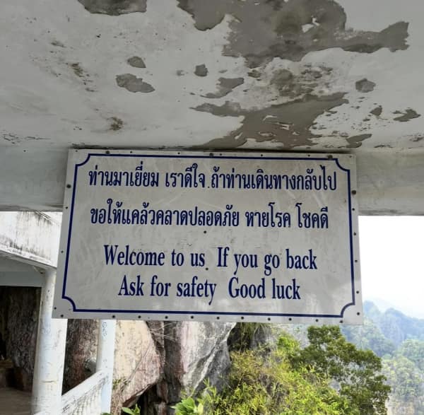 Blue-border sign with Thai script and English lines: “Welcome to us. If you go back Ask for safety Good luck.”