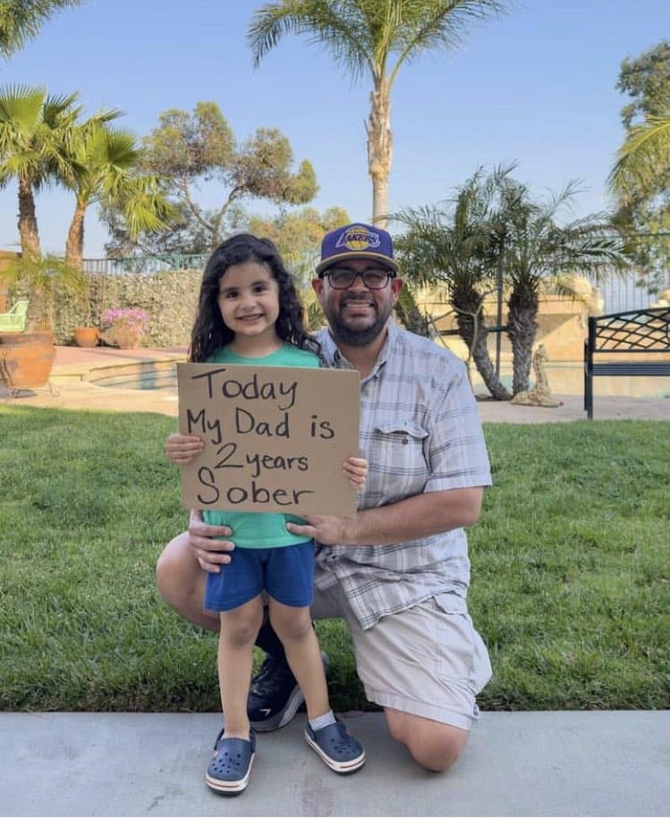 A wholesome meme of a proud daughter holding a sign celebrating her dad's two years of sobriety.