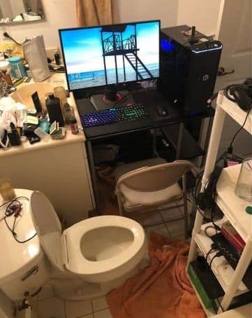 A full gaming PC and monitor setup that has been crammed into a very small and messy bathroom, right next to the toilet.