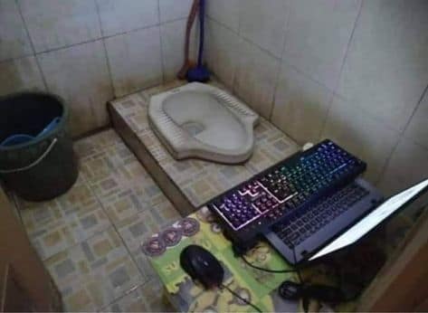 A laptop and an external mechanical gaming keyboard that has been set up on the floor of a bathroom, right next to a squat toilet.