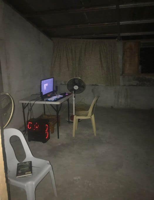 A very minimalist and somewhat depressing gaming computer setup in an empty concrete room with just a single fan and a plastic chair.
