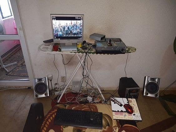 A chaotic and messy computer setup where a wobbly ironing board is being used as a desk, with tangled wires hanging everywhere.