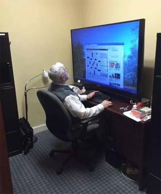 A funny and wholesome photo of an elderly man using a giant, oversized flat-screen television as his computer monitor at a tiny desk.
