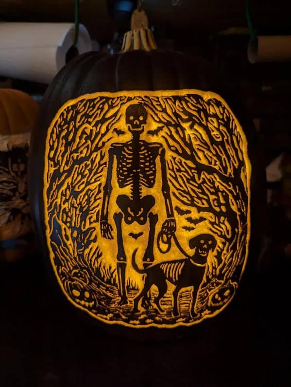A creative jack-o'-lantern featuring a detailed carving of a skeleton walking its skeleton dog in a spooky forest.