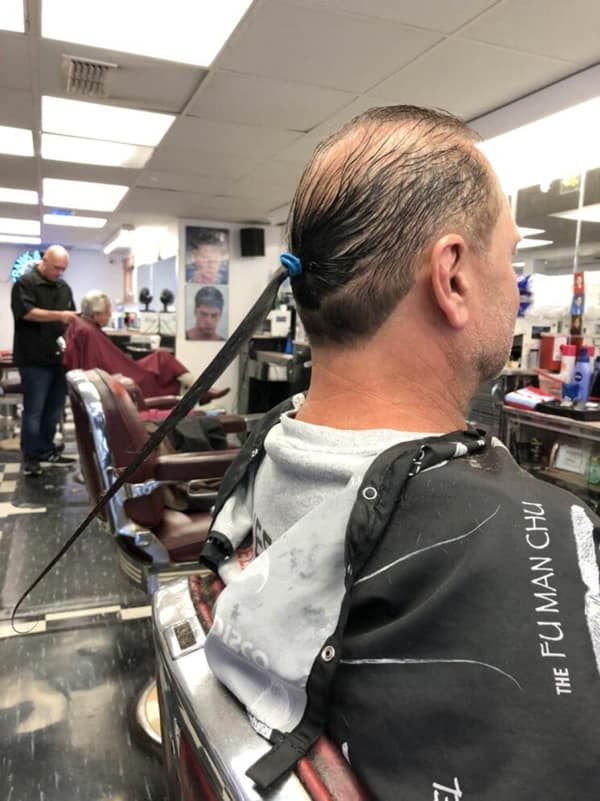 A man in a barbershop with one of the weirdest bad haircuts, a single, long rat tail.
