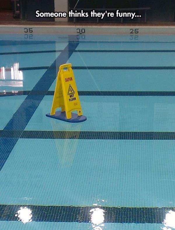 Yellow "Caution Wet Floor" sign floating ironically on a kickboard in a swimming pool.