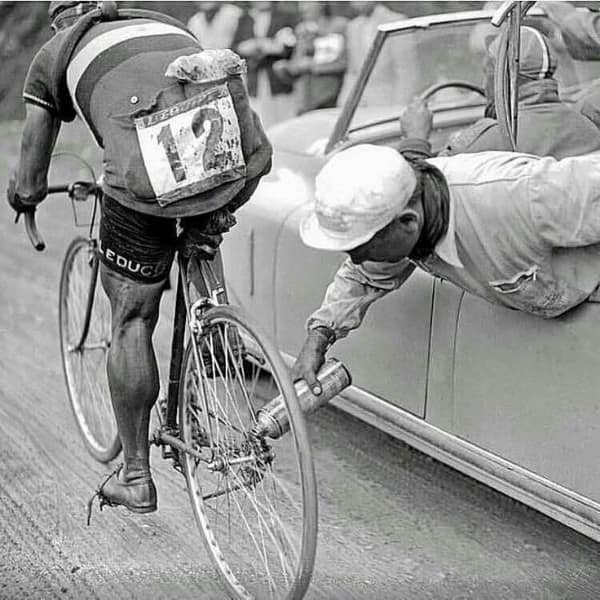 Vintage black and white photo showing a Tour de France cyclist getting his tire lubricated from a moving car.