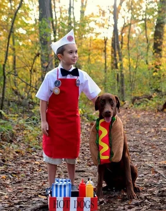 A wholesome dog Halloween costume where a dog is dressed as a hot dog next to a child dressed as a hot dog vendor.