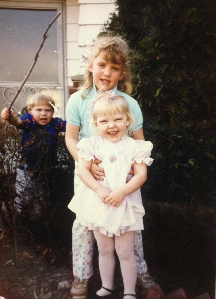 Funny childhood photo of two sisters posing while their brother lurks menacingly behind them holding a stick.