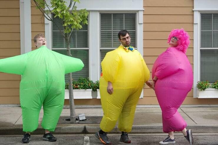 Three family members walking outdoors wearing brightly colored green, yellow, and pink inflatable bodysuits together.