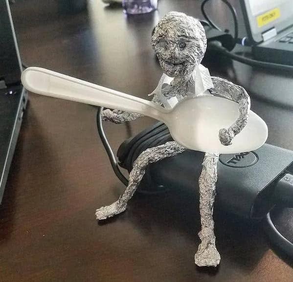 A cute and creative sculpture of a person made from aluminum foil, sitting on a desk and playing a spoon like a guitar.