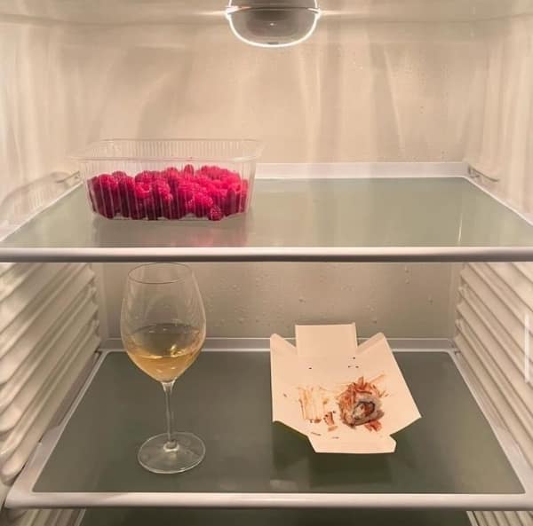 An aesthetic photo of an empty fridge containing only wine, raspberries, and one sushi roll.