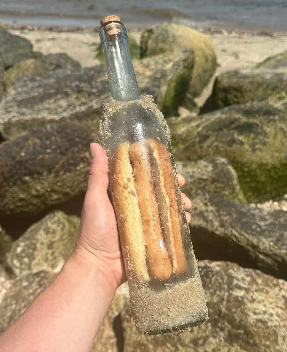 A weird and funny meme of a cursed image showing several hot dogs sealed in a bottle on a beach.