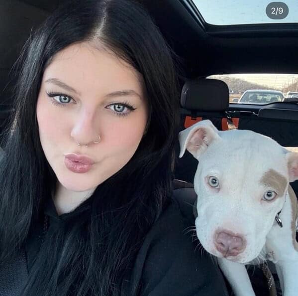 A woman with dark hair and striking blue eyes takes a selfie with her white pitbull puppy with similar blue eyes.