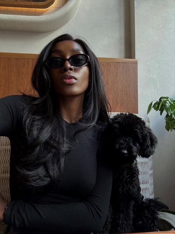 A stylish woman with long black hair and sunglasses sitting next to her small black poodle.