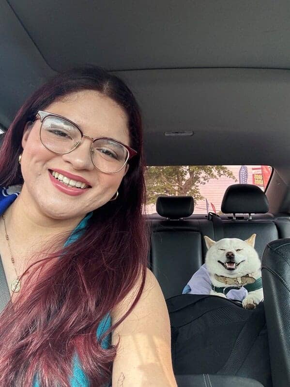 A smiling woman with red hair and glasses taking a car selfie with her happy white Shiba Inu dog.