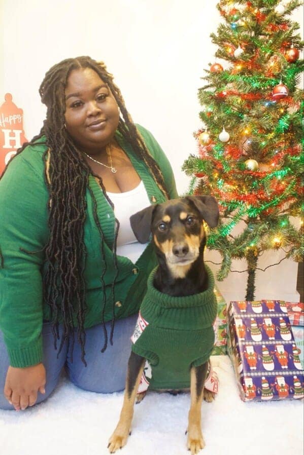 A woman with long locs wearing a green sweater poses with her dog wearing a matching green sweater.