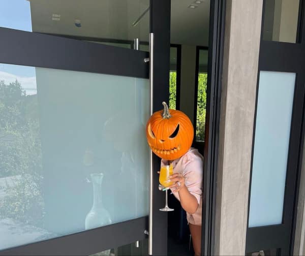A funny picture of a person wearing a carved pumpkin on their head while drinking a mimosa.