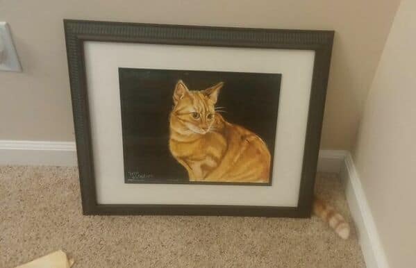 A framed painting of an orange tabby cat, with a real camouflaged cat's tail visible on the floor.