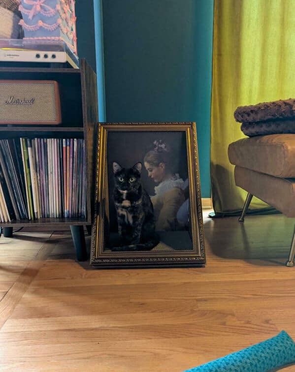 A black cat sitting in front of a framed vintage painting, perfectly camouflaged in the dark hues
