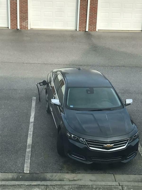 A black car parked in a parking spot with a gas pump nozzle and hose still attached, having driven off.