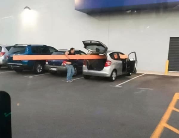 A person in a parking lot failing to load a comically long orange beam into a tiny hatchback.