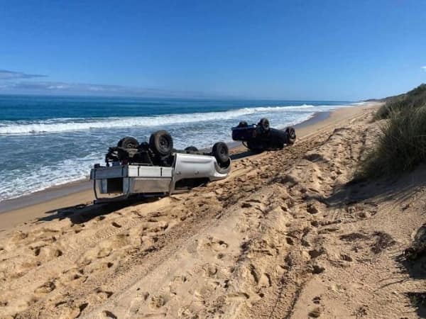 Two off-road 4x4 trucks flipped over and wrecked on a sandy beach, having rolled down a dune.