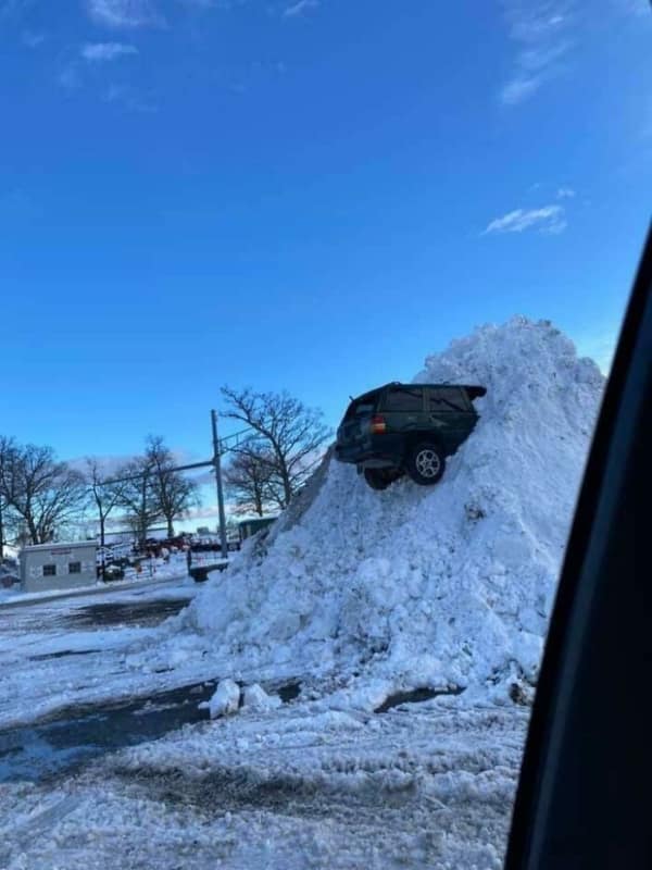 A dark green Jeep or SUV stuck nose-first high up on a giant mountain of plowed snow.