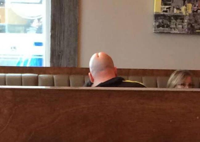 The back of a bald man's head in a restaurant booth, where the crease looks exactly like a butt.