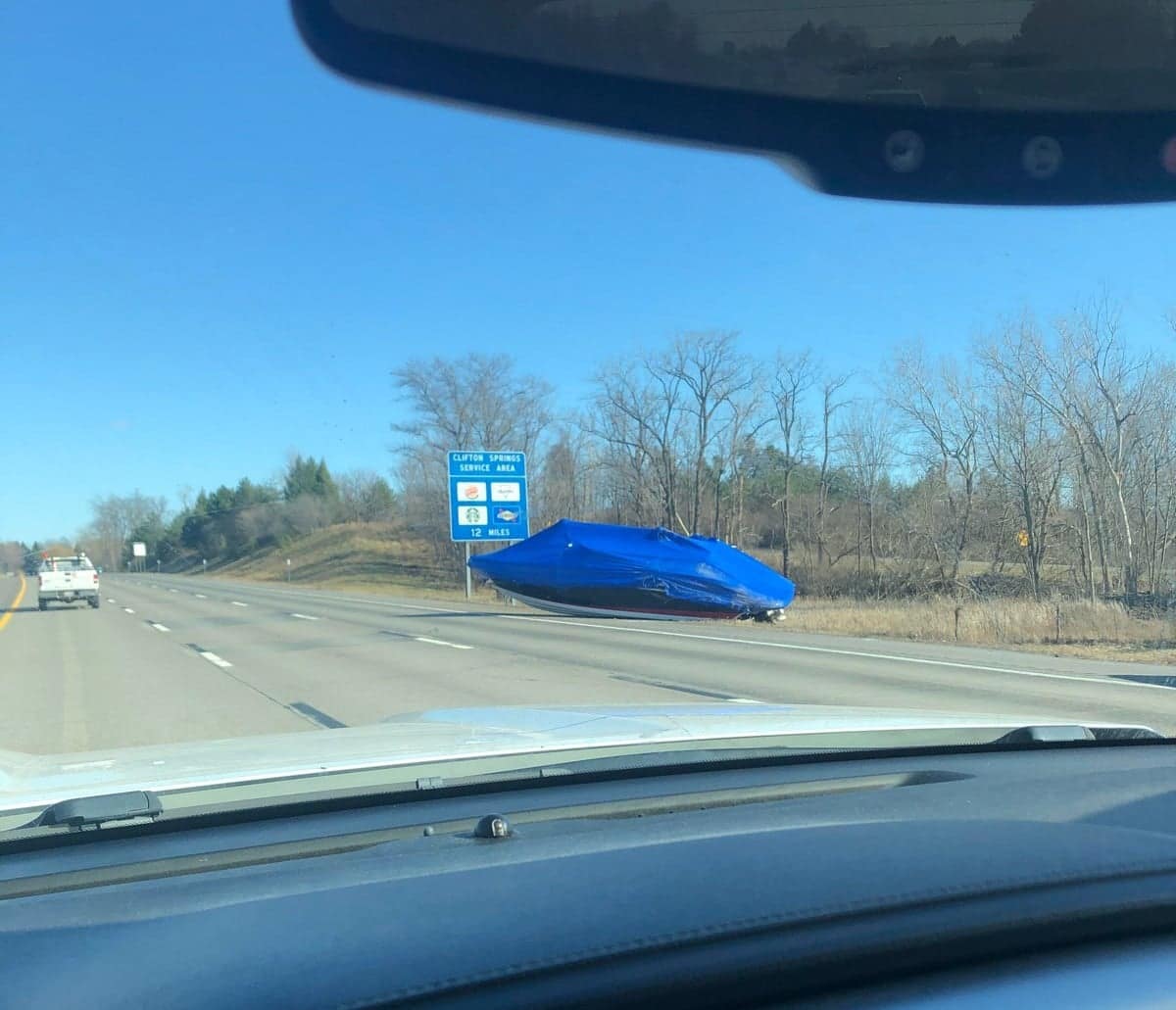 A massive funny fail of a large boat, covered in a blue tarp, that has fallen off its trailer onto a highway median.