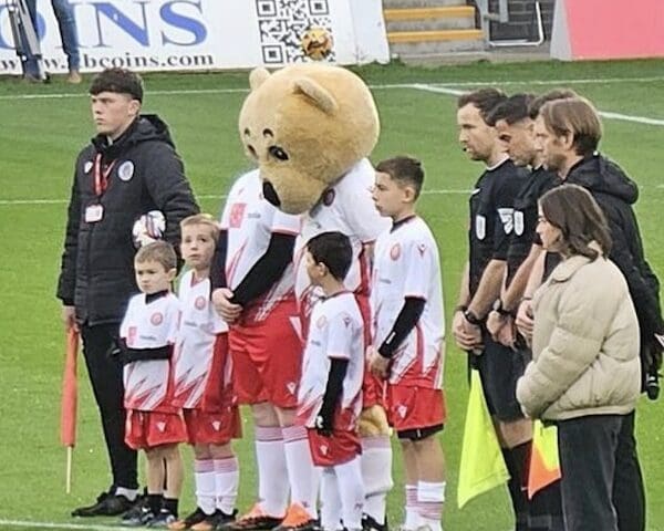A sad-looking teddy bear mascot stands with its head bowed, surrounded by a line of young child players.