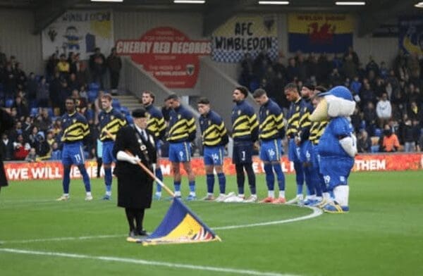 A blue bird mascot stands with its head bowed in a line of football players during a Remembrance Day flag ceremony.