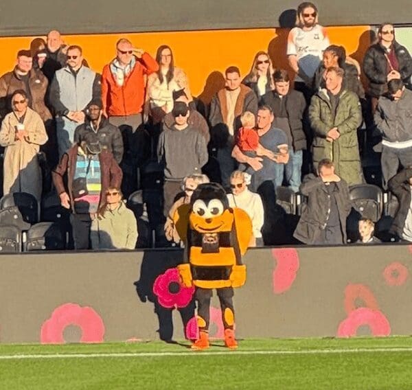 A bee mascot stands solemnly in the crowd holding a large poppy wreath during a moment of silence.