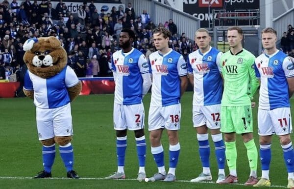 A bear mascot with a blue and white uniform stands with hands behind its back, in line with football players.