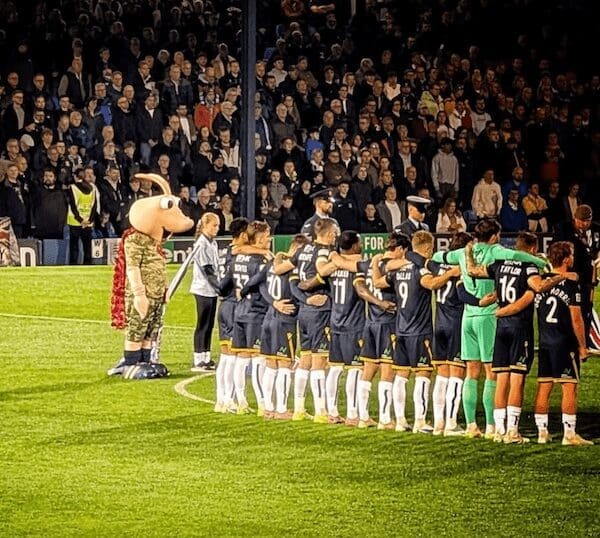 A large pink shrimp mascot wearing a camouflage jacket stands solemnly in front of a football team.