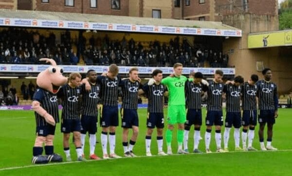 A pink shrimp mascot links arms with a line of football players during a moment of silence on the pitch.