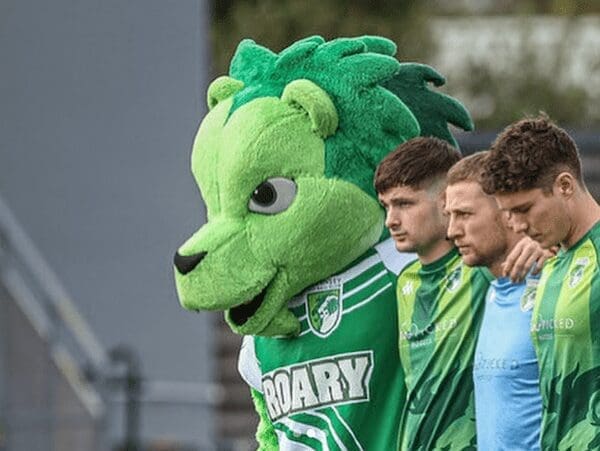 A bright green lion mascot stands in line with football players, looking on with a serious expression.