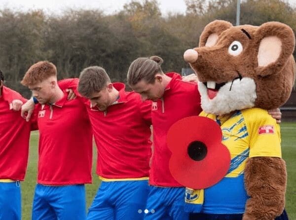A large mouse mascot holding a giant poppy links arms with football players, all bowing their heads in silence.