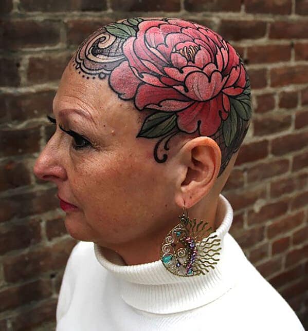 An older, bald woman with a large, beautiful pink peony and filigree tattooed on her head