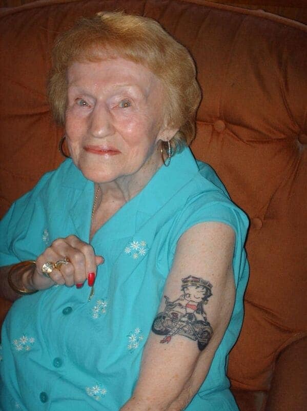 A proud elderly woman with bright red nails, showing off a large Betty Boop on a motorcycle tattoo.