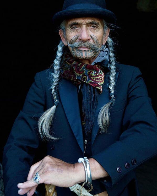A stylish older man with long grey braids, a hat, and a traditional tattoo around his mouth and mustache.