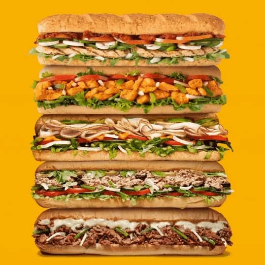 A stack of five delicious-looking Subway footlong sandwiches against a bright yellow background.