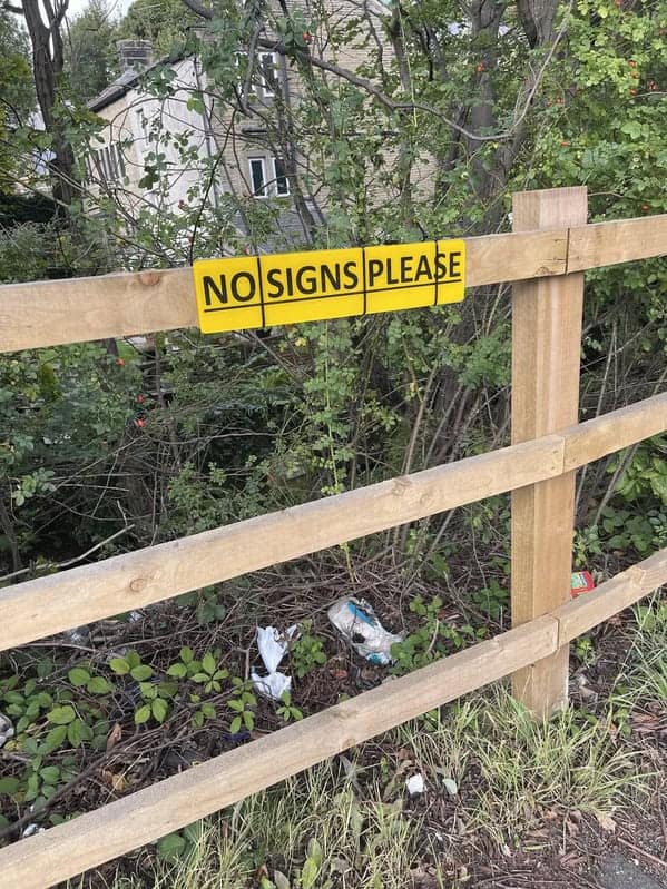 An ironic and funny small yellow sign on a wooden fence that reads, "NO SIGNS PLEASE."