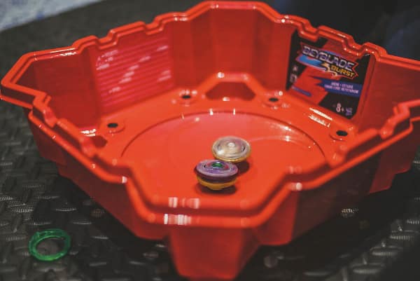 This 90s toy image features a red plastic Beyblade arena with spinning tops.