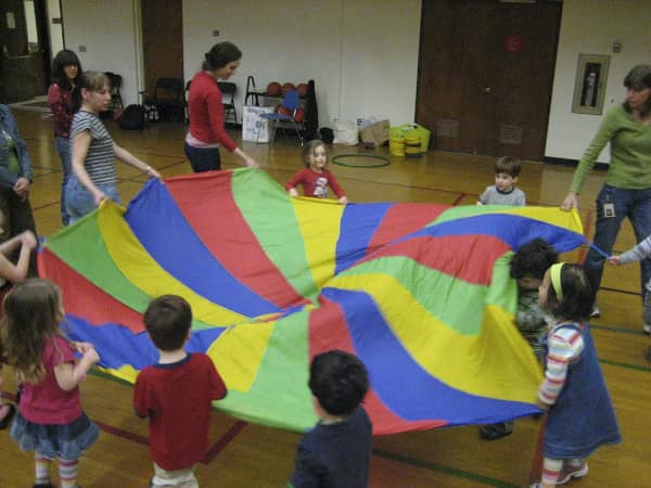 In this 90s toy photo, kids play with a giant multicolor parachute in a gym.